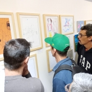 Uma parede branca com quadros expostos. Pessoas olham os trabalhos expostos.