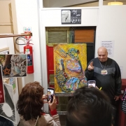 Um quadro exposto no cavalete, com texturas em 3 dimensões, colorido, com prevalência de tons amarelos. Um homem branco, calvo, vestindo um moletom preto, faz sinal com a mão e sorri para a foto.