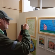 Um homem idoso fotografa um quadro com o celular. Ele é branco, usa um boné e um óculos de grau e veste um casaco verde escuro.