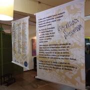 Banners com a identidade visual da exposição estão suspensos. Eles são brancos, com tons de bege num desenho abstrato e as letras dos textos escritos são em cor preta. 