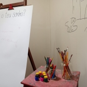 Um quadro branco com a frase "Qual teu sonho?" escrito à mão com caneta preta no lado esquerdo da foto. No lado direito, uma mesinha está colocada. Em cima dela estão pinceis, tintas e lápis de cor postos.