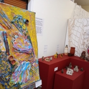 Um quadro com texturas em 3 dimensões, colorido, com a cor prevalente amarelo está à esquerda. Um móvel de madeira vermelho apoiando peças de cerâmica está do lado direito. A parede ao fundo é branca. 