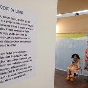 Uma parede branca no lado esquerdo onde está colado um cartaz igualmente branco com um texto em letras pretas. No lado direito da imagem dois bonecos de pano estão posicionados.
