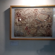 Um quadro com uma obra em bordado está disposto em uma parede branca. O quadro é abstrato, mas retrata um ambiente rural com a figura prevalente de patos e galinhas.