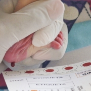 Uma profissional de saúde, com luvas descartáveis, realizando a coleta de sangue do calcanhar de um recém-nascido para o Teste do Pezinho. O bebê está deitado, e a profissional utiliza uma lanceta para realizar a punção, enquanto segura o pé do bebê com cuidado. A cena transmite um ambiente clínico limpo e organizado, destacando a atenção e o cuidado dedicados à saúde neonatal