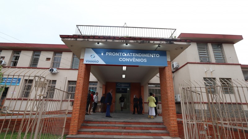 A imagem mostra a entrada de um prédio de atendimento médico. Na parte superior da entrada, há uma placa azul com os dizeres "PRONTO ATENDIMENTO CONVÊNIOS" e o logotipo da Prefeitura de Viamão. A fachada do prédio é composta por tijolos vermelhos e paredes brancas. Há janelas com grades e um portão de ferro na frente. Algumas pessoas estão posicionadas próximas à entrada do prédio.