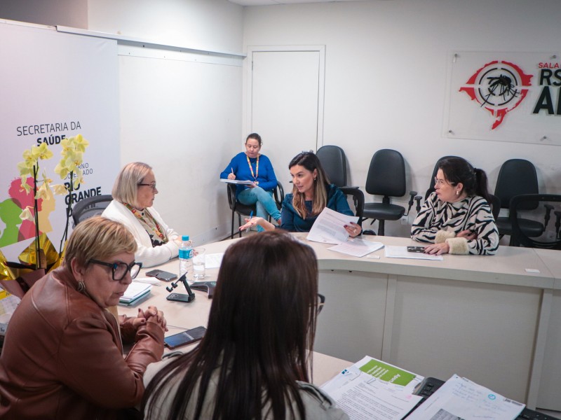 A imagem retrata uma reunião em andamento em uma sala de conferências. Seis pessoas estão sentadas ao redor de uma mesa retangular. A mesa está coberta com papéis, garrafas de água e materiais de escritório. Na parede à esquerda, há um banner com os dizeres "SECRETARIA DA SAÚDE RIO GRANDE DO SUL". Na parede à direita, vê-se um logotipo acompanhado do texto "SAÚDE RS". Ao fundo da sala, há várias cadeiras vazias encostadas na parede.