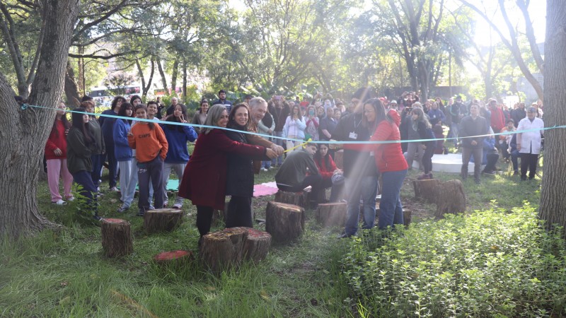 O jardim com várias plantas e árvores está repleto de participantes do evento. Autoridades que participaram da inauguração seguram fita e sorriem.