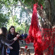 Duas meninas vestindo roupas pretas tocam instalação feita com fio de crochê vermelho pendurado numa árvore.