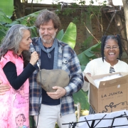 Da esquerda pra direita, Simone fala ao microfone, acompanhada do representante do Jardim Botânico e da artista Solange Luciano. Simone é uma mulher branca, de cabelos grisalhos longos. Ela veste uma blusa preta e uma veste falante na cor rosa. O homem ao seu lado é branco, de cabelos loiros e barba grisalha. Ele veste uma camisa de flanela em tons azuis. Solange é uma mulher preta, sorridente. Ela vesta uma roupa listrada nas cores preto e branco e um casaco na cor creme. Ela segura uma caixa de papelão.