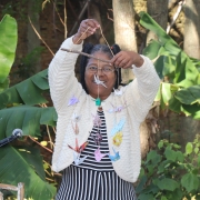 Solange é uma mulher preta, sorridente. Ela veste uma roupa listrada nas cores preta e branca e um casaco cor creme. Ela segura um móbile artesanal feito com dobraduras de papel colorido e barbante.
