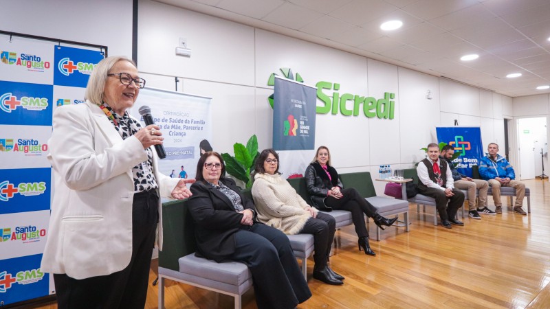 secretária Arita de casaco branco ao microfone com outras seis pessoas sentadas em poltronas ao seu lado
