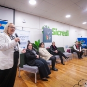 secretária Arita de casaco branco ao microfone  com outras seis pessoas sentadas em poltronas ao seu lado 