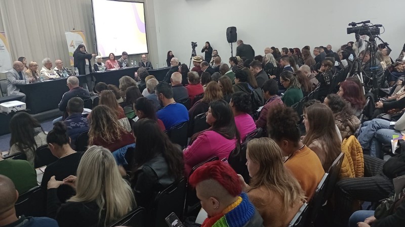 A imagem mostra uma sala de conferências cheia de pessoas sentadas, assistindo a uma apresentação. Na parte frontal da sala, há uma mesa longa com várias pessoas sentadas atrás dela. Uma tela grande ao fundo exibe uma projeção. Algumas pessoas na plateia estão tirando fotos ou fazendo anotações. A sala está bem iluminada, com equipamentos de gravação e câmeras posicionados ao redor para registrar o evento.