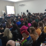 A imagem mostra uma sala de conferências cheia de pessoas sentadas, assistindo a uma apresentação. Na parte frontal da sala, há uma mesa longa com várias pessoas sentadas atrás dela. Uma tela grande ao fundo exibe uma projeção. Algumas pessoas na plateia estão tirando fotos ou fazendo anotações. A sala está bem iluminada, com equipamentos de gravação e câmeras posicionados ao redor para registrar o evento.