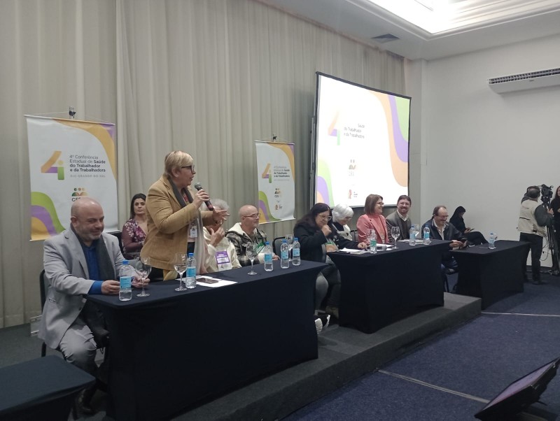 A imagem mostra uma conferência ou mesa de discussão com várias pessoas sentadas atrás de uma mesa longa sobre um palco. Ana Costa está falando ao microfone. Em ambos os lados do palco, há banners com o texto “4ª Conferência Estadual de Saúde das Trabalhadoras e dos Trabalhadores” e logotipos na parte inferior. Ao fundo, uma tela grande exibe o mesmo texto e logotipos. Sobre a mesa, há garrafas de água e microfones, indicando que se trata de um evento formal e organizado.