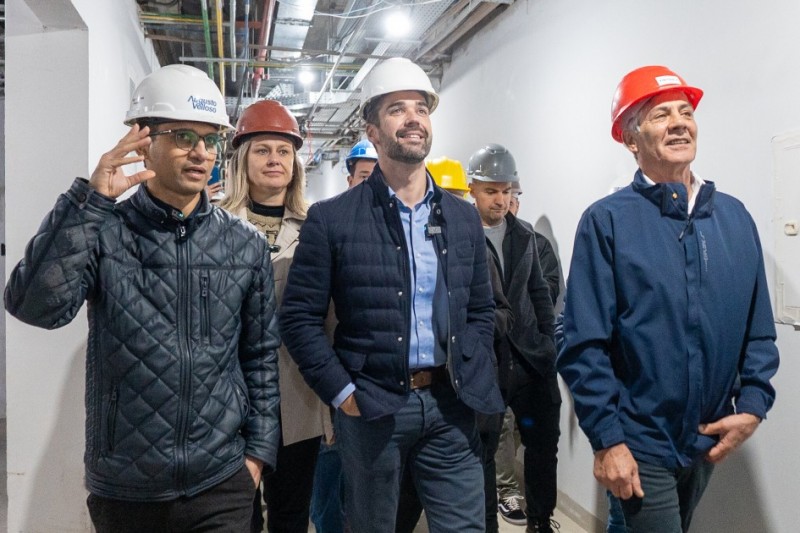 Seis pessoas - entre elas o governador Eduardo Leite e o prefeito de Pelotas, Fernando Marroni, caminham por um corredor iluminado, todas usando capacetes de segurança de cores variadas — branco, vermelho, cinza e azul. Estão vestidas com roupas casuais e jaquetas.