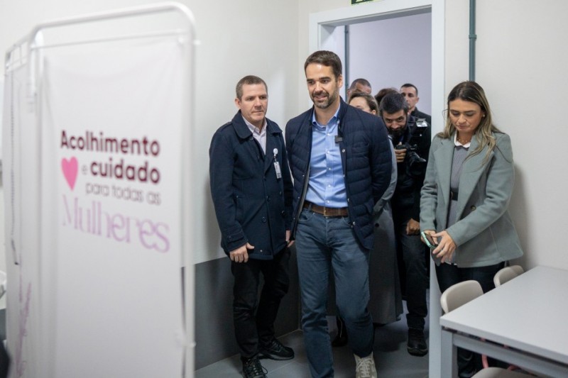 Grupo de pessoas caminha por um corredor de uma instalação médica. À esquerda, há uma divisória com a frase 'Acolhimento e cuidado para todas as Mulheres' e um ícone de coração rosa.