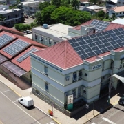 Vista aérea de um complexo de edifícios com painéis solares instalados nos telhados. Os prédios estão cercados por ruas e veículos estacionados. O edifício principal exibe uma placa com os dizeres 'ACHA- Associação Comunitária Hospital de Aratiba