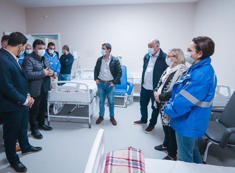 A imagem mostra um grupo de sete pessoas em pé dentro de um quarto de hospital. O ambiente contém duas camas hospitalares, sendo que uma delas está coberta com um cobertor xadrez. Também há duas cadeiras azuis e uma cadeira cinza no local. As pessoas estão vestidas com roupas casuais; algumas usam jaquetas azuis do SUS com detalhes refletivos. Eles estão usando máscara por estar em ambiente hospitalar