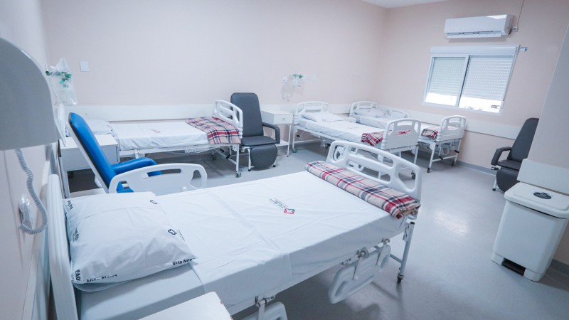 A imagem retrata um quarto de hospital com seis camas brancas, todas equipadas com grades laterais e travesseiros. Três dessas camas têm cobertores xadrez dobrados aos pés. Entre as camas, há duas cadeiras pretas e uma cadeira azul. Na parede do fundo, vê-se uma janela com persianas fechadas e, acima dela, um aparelho de ar-condicionado. O chão é de cor cinza claro e as paredes são bege claro. À direita da imagem, próximo à porta, há uma lixeira branca.