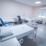 A imagem retrata um quarto de hospital com seis camas brancas, todas equipadas com grades laterais e travesseiros. Três dessas camas têm cobertores xadrez dobrados aos pés. Entre as camas, há duas cadeiras pretas e uma cadeira azul. Na parede do fundo, vê-se uma janela com persianas fechadas e, acima dela, um aparelho de ar-condicionado. O chão é de cor cinza claro e as paredes são bege claro. À direita da imagem, próximo à porta, há uma lixeira branca.