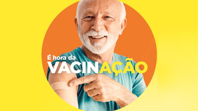 um idoso branco e sorridente mostra o braço vacinado o fundo é amarelo e laranja e em letras brancas e amarelas o slogan - É hora da vacinação