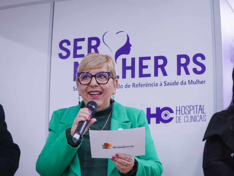 Ana Costa segura um microfone e um papel enquanto fala em um evento. Ela veste um blazer verde sobre uma blusa preta de gola alta, usa um colar dourado e brincos. Ao fundo, há um painel com os dizeres "SER MULHER RS Serviço Estadual de Referência à Saúde da Mulher" e o logotipo do Hospital de Clínicas (HC).