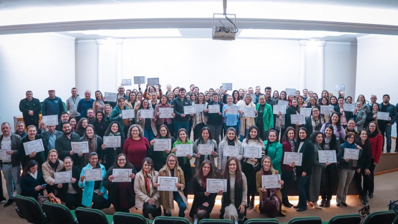 m grande grupo de pessoas está reunido em um auditório, distribuído em três fileiras e posando para uma foto coletiva. A maioria segura certificados nas mãos. O ambiente é bem iluminado, com cadeiras verdes visíveis na parte inferior da imagem, indicando que a foto foi tirada de frente para o palco.