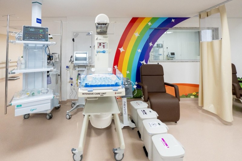 Fotografia de uma unidade de terapia intensiva neonatal. No centro da imagem, há uma incubadora com a inscrição 'INTENSIVE CARE UNIT' e a marca 'Fanem'. À esquerda, há um suporte com equipamentos médicos, como monitores e tubos. À direita da incubadora, vê-se uma poltrona marrom para acompanhantes e três lixeiras brancas identificadas para descarte de resíduos. Ao fundo, um mural colorido com arco-íris e estrelas decora a parede. Uma cortina bege à direita pode ser usada para garantir privacidade.