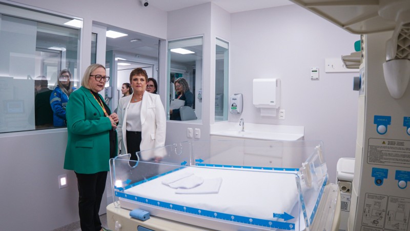 A imagem mostra uma sala de hospital, possivelmente uma unidade neonatal, com um ber&ccedil;o aquecido em primeiro plano. Duas pessoas est&atilde;o conversando ao lado do ber&ccedil;o. Arita Bergmann est&aacute; vestida com um blazer verde e a outra com um blazer branco. Ao fundo, h&aacute; mais pessoas observando atrav&eacute;s de uma janela de vidro. A sala &eacute; bem iluminada e possui equipamentos m&eacute;dicos nas paredes e no teto.