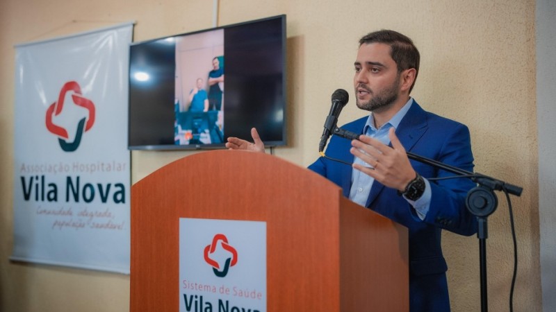 A imagem mostra o vice-governador Gabriel Souza vestindo um terno azul e falando em um p&uacute;lpito de madeira. No p&uacute;lpito, h&aacute; um logotipo e o texto "Sistema de Sa&uacute;de Vila Nova". &Agrave; esquerda, uma faixa branca exibe o mesmo logotipo e a frase: "Associa&ccedil;&atilde;o Hospitalar Vila Nova - inova&ccedil;&atilde;o, seguran&ccedil;a, prop&oacute;sitos saud&aacute;veis". Ao fundo, uma televis&atilde;o mostra a imagem de duas pessoas.