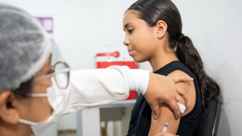 Adolescente recebe vacina no bra&ccedil;o esquerdo, aplicada por profissional de sa&uacute;de com jaleco branco e luvas, em ambiente cl&iacute;nico.