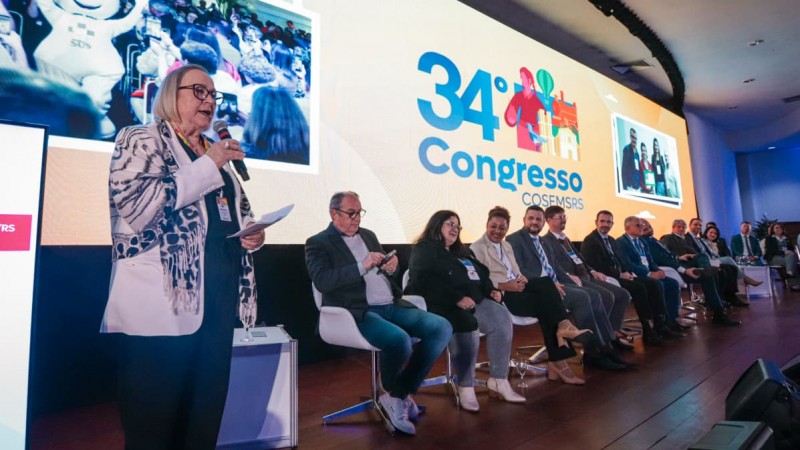 A imagem mostra um evento formal realizado em um palco. H&aacute; uma longa fileira de cadeiras ocupadas por v&aacute;rias pessoas sentadas, autoridades do evento. &Agrave; esquerda da imagem, Arita Bergmann est&aacute; em p&eacute;, segurando um microfone e alguns pap&eacute;is, discursando para o p&uacute;blico.

Ao fundo, destaca-se um grande painel digital com o texto "34&ordm; Congresso COSEMS/RS", acompanhado de gr&aacute;ficos coloridos. Tamb&eacute;m h&aacute; duas telas menores que exibem imagens relacionadas ao evento.