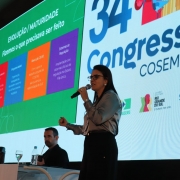 Diretora do Dgae, Lisiane Fagundes fala ao microfone tendo ao fundo um painel do 34º Congresso do Cosems 