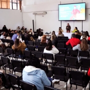 Em um auditório com as paredes brancas e cadeiras na cor preta, uma plateia assiste a uma palestra. Ao centro, uma mulher de cabelos pretos na altura dos ombros, vestindo roupa preta, segura o microfone. Um telão com projeção está atrás e acima dela.
