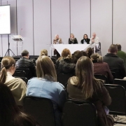 a imagem é de um ambiente formal de conferência ou seminário, com quatro palestrantes em um palco, uma plateia atenta e uma tela de apresentação ao lado.