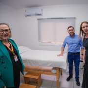 A imagem mostra quatro pessoas em pé dentro de uma sala com uma maca coberta por um lençol branco. A sala tem paredes brancas, um ar-condicionado instalado na parede e uma janela com persianas.