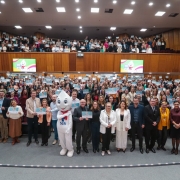 Entrega do Selo Município Amigo da Vacina 2024. A imagem mostra um grande grupo de pessoas reunidas em um auditório, segurando certificados. Há um mascote do Zé Gotinha. Duas telas exibem a mesma imagem do mascote acompanhada de texto.