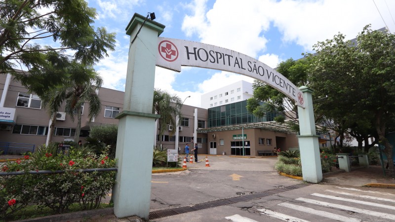 A imagem mostra a entrada do Hospital S&atilde;o Vicente. H&aacute; um arco com a inscri&ccedil;&atilde;o "HOSPITAL S&Atilde;O VICENTE" e um s&iacute;mbolo m&eacute;dico em vermelho. O hospital &eacute; um edif&iacute;cio de v&aacute;rios andares, com uma fachada bege e janelas brancas. H&aacute; &aacute;rvores e plantas ao redor da entrada, bem como uma faixa de pedestres em primeiro plano.