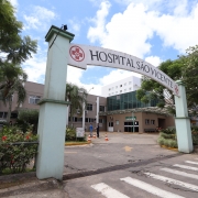 A imagem mostra a entrada do Hospital S&atilde;o Vicente. H&aacute; um arco com a inscri&ccedil;&atilde;o "HOSPITAL S&Atilde;O VICENTE" e um s&iacute;mbolo m&eacute;dico em vermelho. O hospital &eacute; um edif&iacute;cio de v&aacute;rios andares, com uma fachada bege e janelas brancas. H&aacute; &aacute;rvores e plantas ao redor da entrada, bem como uma faixa de pedestres em primeiro plano.