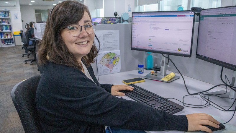 A imagem mostra uma pessoa sentada em uma mesa de escrit&oacute;rio, trabalhando em um computador com dois monitores. H&aacute; diversos itens sobre a mesa, como um smartphone, post-its e pequenos objetos. Ao fundo, v&ecirc;-se outras pessoas em seus postos de trabalho e estantes com livros.