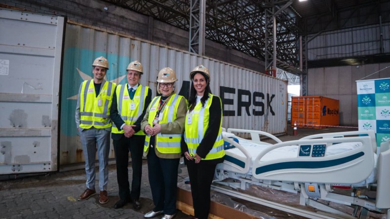 A imagem mostra quatro pessoas com coletes e capacetes de seguran&ccedil;a em ambiente industrial, ao lado de uma cama hospitalar moderna. Ao fundo, h&aacute; cont&ecirc;ineres de carga, incluindo um com a marca Maersk. Arita Bergmann est&aacute; presente na foto.