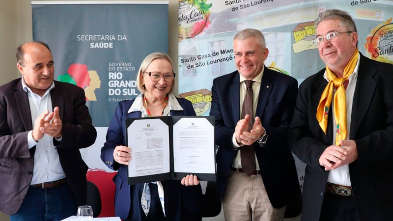 A imagem mostra Arita Bergman segurando um documento durante uma cerim&ocirc;nia oficial, acompanhada por tr&ecirc;s homens que aplaudem. Ao fundo, h&aacute; banners da Secretaria da Sa&uacute;de, do Governo do Estado do RS e da Santa Casa de Miseric&oacute;rdia de S&atilde;o Louren&ccedil;o do Sul.