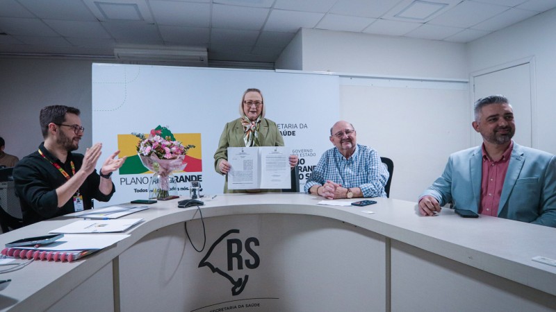 Ao centro da foto a secret&aacute;ria segura uma pasta com os documentos assinados. Ela &eacute; uma mulher branca, com cabelos loiros na altura dos ombros e veste uma roupa verde. No lado esquerdo um homem branco vestindo uma camisa preta bate palmas. Ao lado direito da mesa dois homens vestindo roupas azul claro olham para a frente e sorriem. 