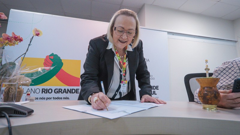 Arita assina um documento em cima de uma mesa. Ela veste uma camisa branca um blazer preto. Ao lado direito dela est&aacute; uma cuia de chimarr&atilde;o.