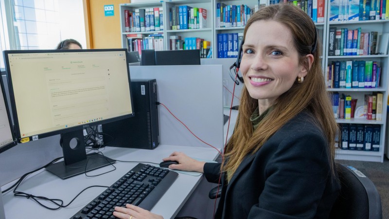 A imagem mostra uma pessoa sentada em frente a um computador, sorrindo para a c&acirc;mera, em um ambiente que parece ser uma biblioteca ou escrit&oacute;rio. Ao fundo, h&aacute; estantes com livros organizados, e o monitor exibe uma p&aacute;gina da web com texto n&atilde;o leg&iacute;vel.