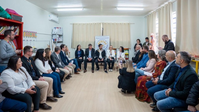 Uma sala de aula em tons claros est&aacute; organizada com as autoridades sentadas em forma de U. O governador est&aacute; ao centro da foto, ao lado da secret&aacute;ria Arita Bergmann.