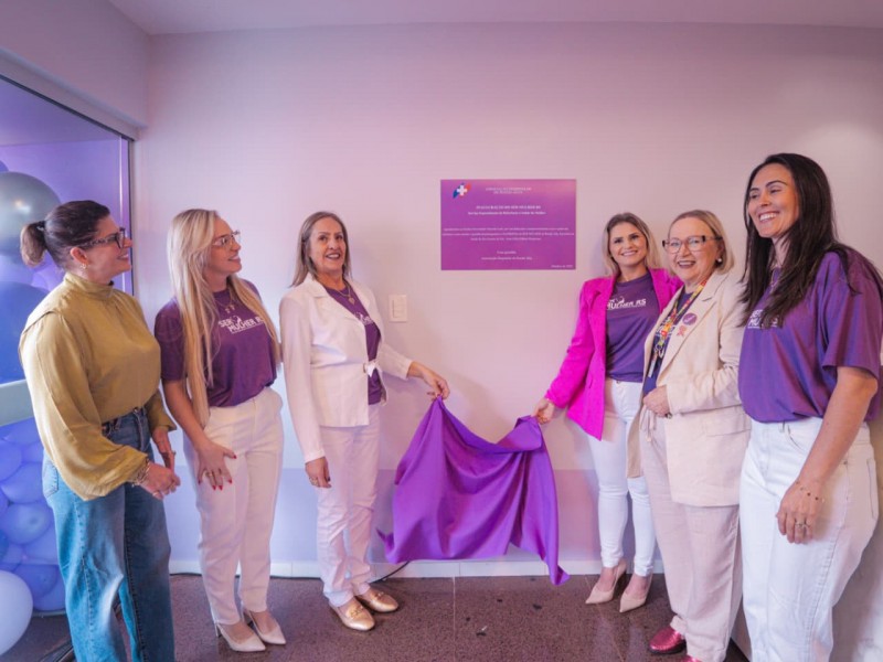 Seis pessoas estão posicionadas em frente a uma parede clara, onde há uma placa roxa fixada. Duas delas seguram um tecido roxo parcialmente aberto, indicando um momento de inauguração ou descerramento da placa. As pessoas vestem roupas predominantemente em tons de roxo e branco, com algumas peças em cores diferentes, como uma blusa amarela. À esquerda, há uma janela com balões decorativos em tons de roxo e branco. O ambiente é interno, com piso brilhante.