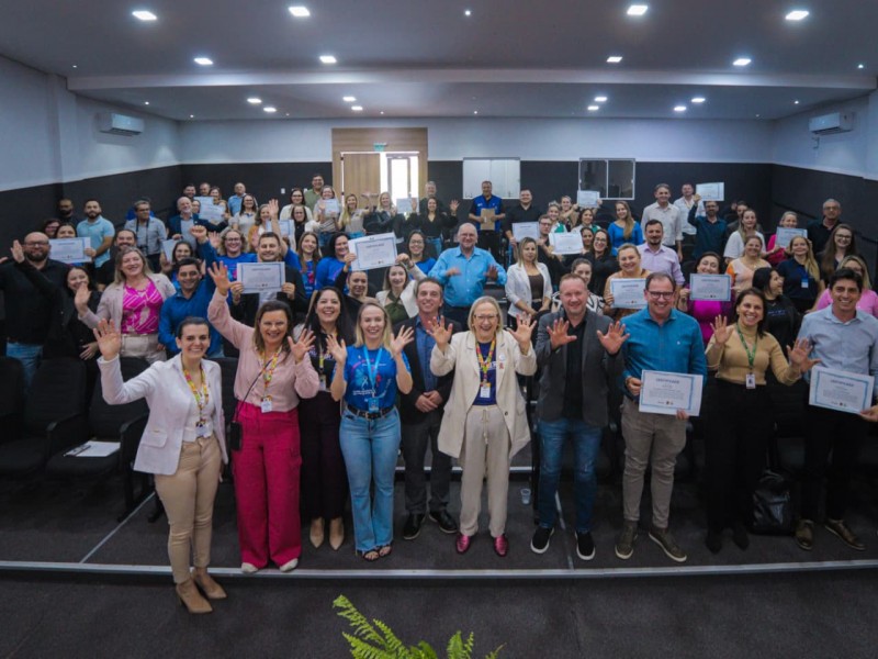 A imagem mostra um auditório com várias pessoas reunidas para um evento formal. No centro e nas primeiras fileiras, participantes seguram certificados, indicando que se trata da entrega do certificado “Equipe Amiga da Mãe, Parceria e Criança 2024”. O ambiente é iluminado, com cadeiras organizadas em fileiras e uma mesa ou espaço livre à frente. Ao fundo, há mais pessoas em pé, também exibindo certificados, sugerindo uma cerimônia coletiva de reconhecimento. A atmosfera geral é de celebração e valorização, com destaque para a participação ativa dos presentes.